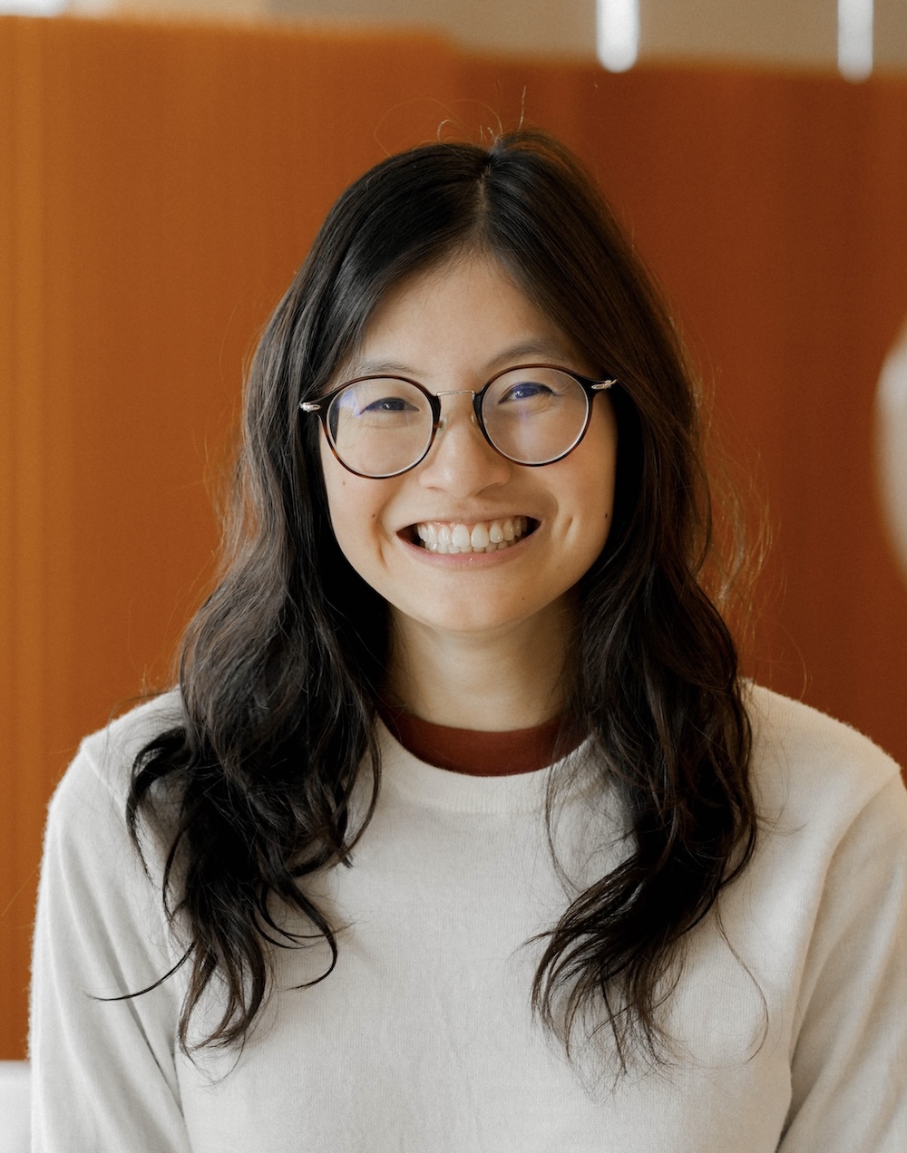 Smiling woman with shoulder-length black hair, wearing glasses and a striped shirt, her arms folded